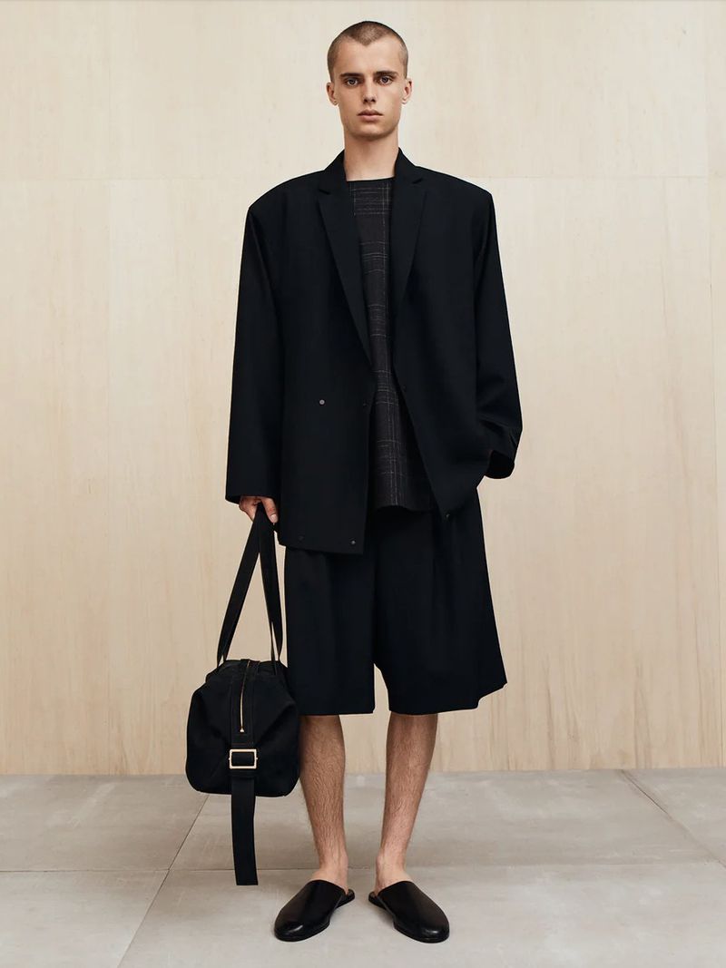 A young adult male in a black blazer, shorts, and slides poses with a black duffel bag against a light tan backdrop. Fashion editorial.