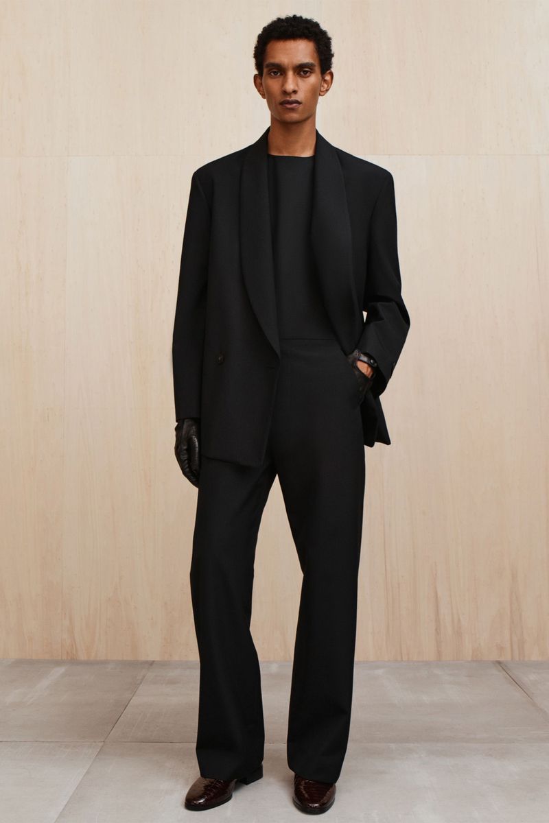 A full-length shot of an adult male posing in a black suit with black gloves and brown shoes in front of a neutral background.