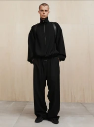 Model wearing an all-black high-neck pullover and wide-leg trousers against a light wood panel wall.