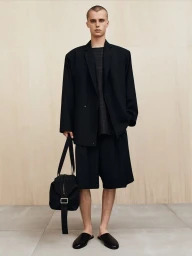Model wearing an oversized black blazer and matching shorts holding a black bag against a light wood panel wall.