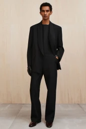 Model wearing a black tailored suit with shawl lapels and black leather gloves standing in front of a light wood paneled wall.