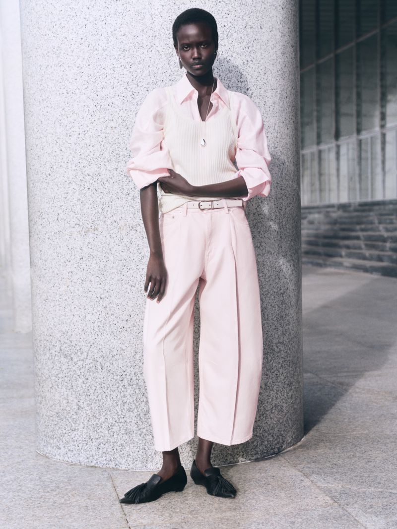 Model standing next to a large concrete pillar wearing a light pink shirt, cream knit vest, and matching trousers.