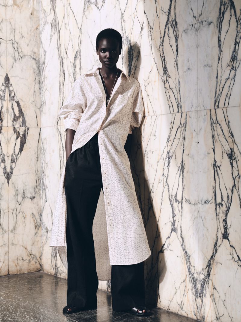 Indoor shot of a Black woman fashion model wearing a long, light-colored shirt and black slacks leaning against a marble wall.