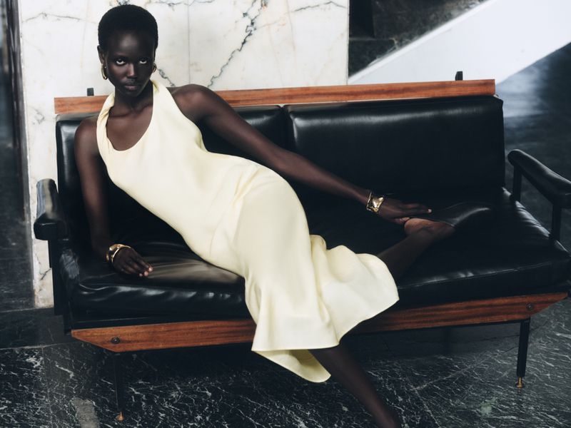 Elegant dark-skinned woman in a cream-colored halter dress reclining on a black leather sofa against a marble wall backdrop.