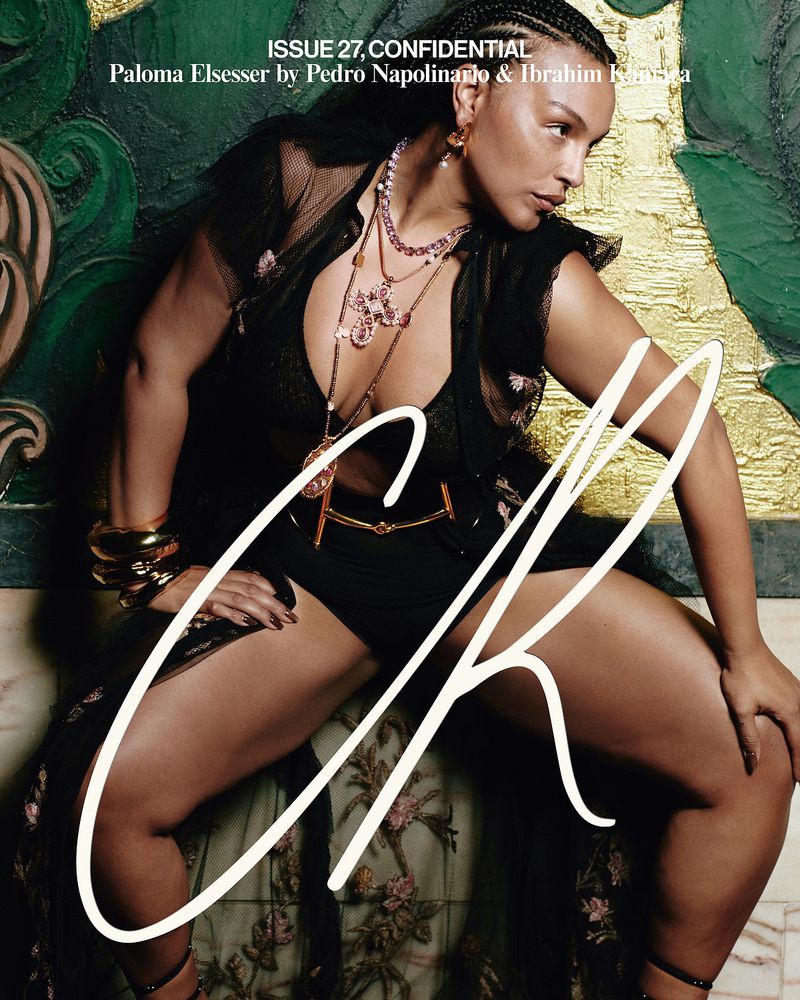 Paloma Elsesser poses in a black outfit with layered necklaces, gold bracelets and anklets on a painted decorative surface for a magazine cover.