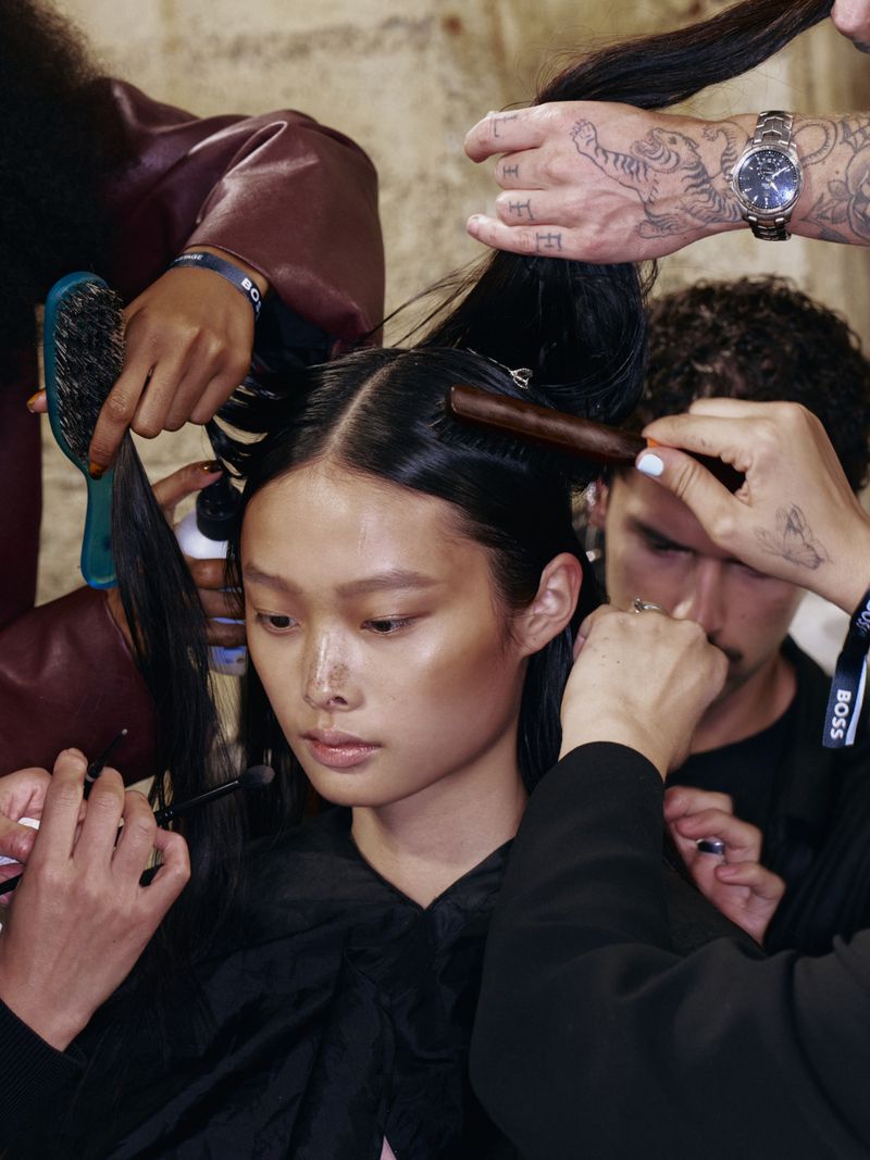 Backstage image of an Asian female model having her hair styled by multiple stylists, highlighting beauty, fashion, and backstage preparations.