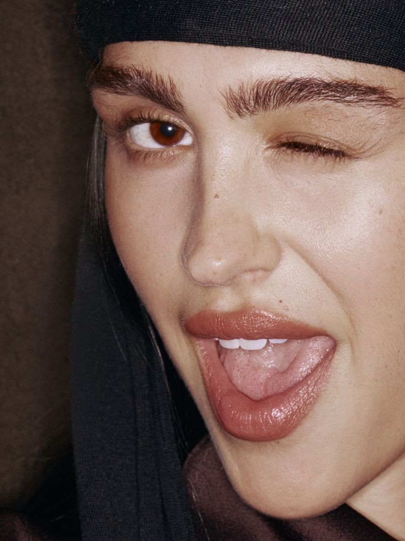Close-up portrait of an adult woman with an open mouth and one eye winking, wearing a black cap and visible brown top.