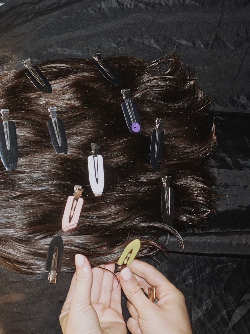 Hair styling with visible hands, dark hair, and metal clips on a black satin background, viewed from overhead in a beauty concept image.