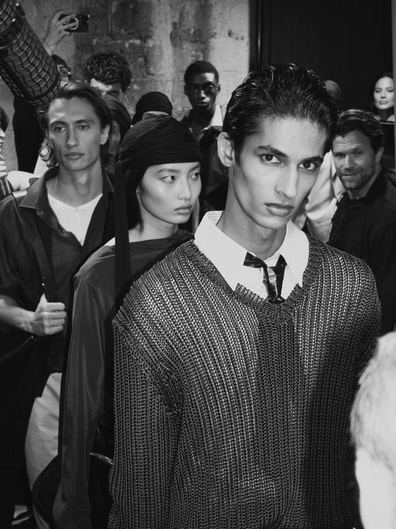 Backstage black-and-white fashion photograph: a stylish group of male and female models in knitwear, with concrete wall backdrop.