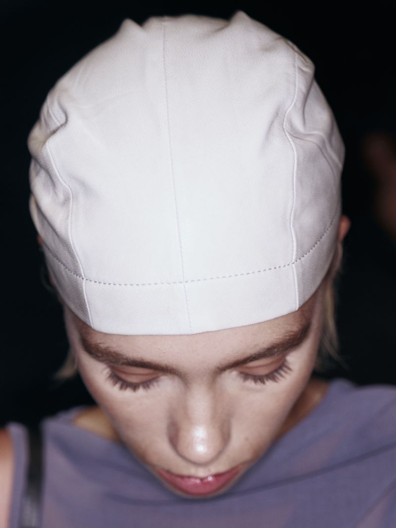 Eye-level close-up of a fair-skinned adult female wearing a close-fitting white cap, looking down in an indoor setting with dark background.
