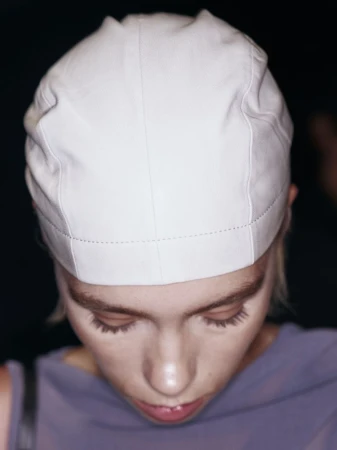 Eye-level close-up of a fair-skinned adult female wearing a close-fitting white cap, looking down in an indoor setting with dark background.