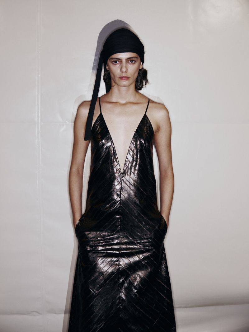 Woman in black dress with head wrap stands in front of neutral wall in studio with soft lighting, fashion editorial photography.
