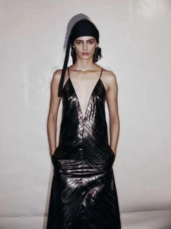 Woman in black dress with head wrap stands in front of neutral wall in studio with soft lighting, fashion editorial photography.