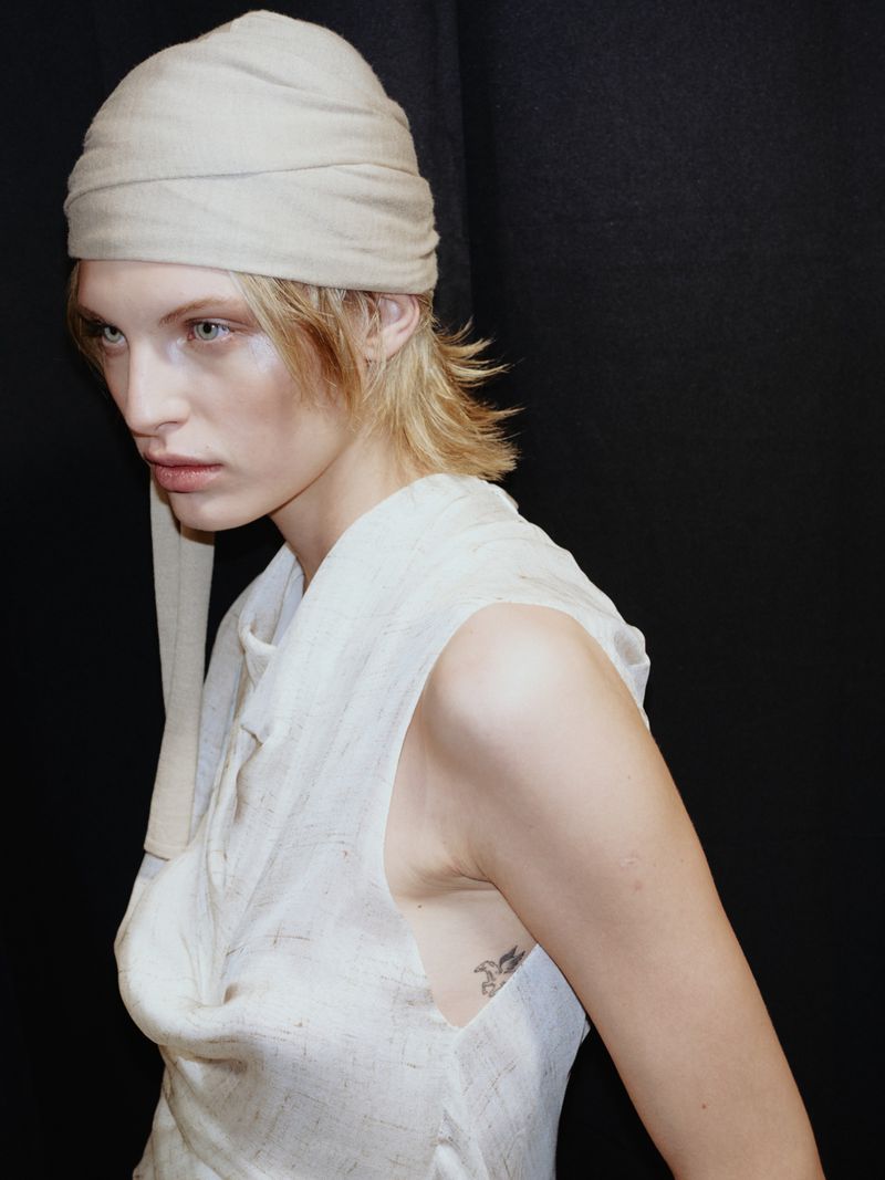 Close-up studio portrait of a woman with blonde hair wearing a linen headwrap and sleeveless top against a dark background.