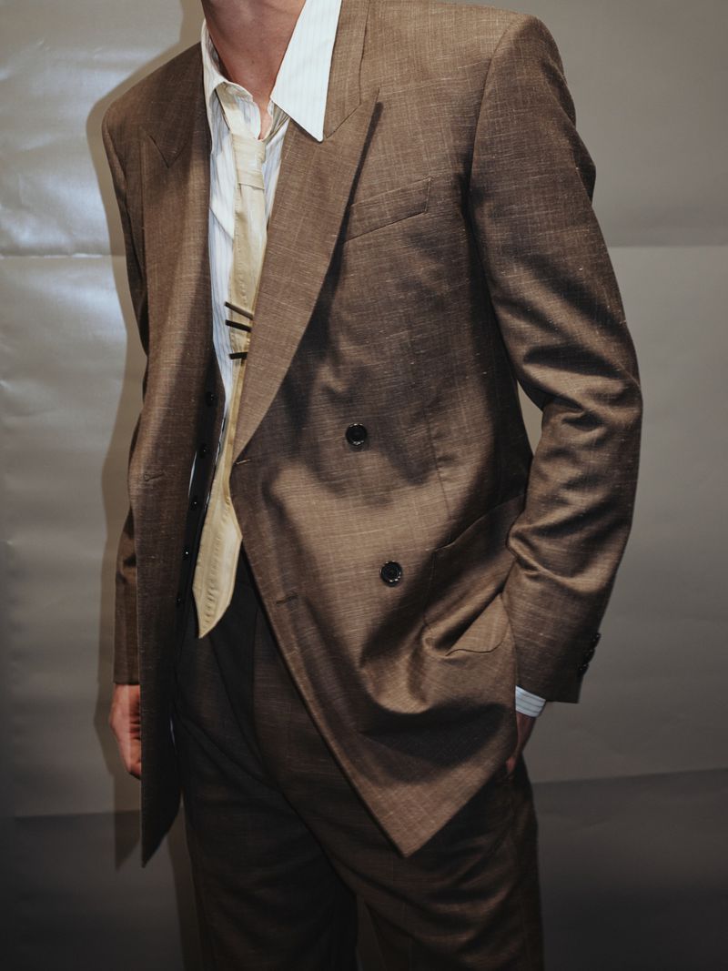 Man in a brown double-breasted suit with beige tie and white shirt, indoors with a neutral background, in a fashion or editorial context.