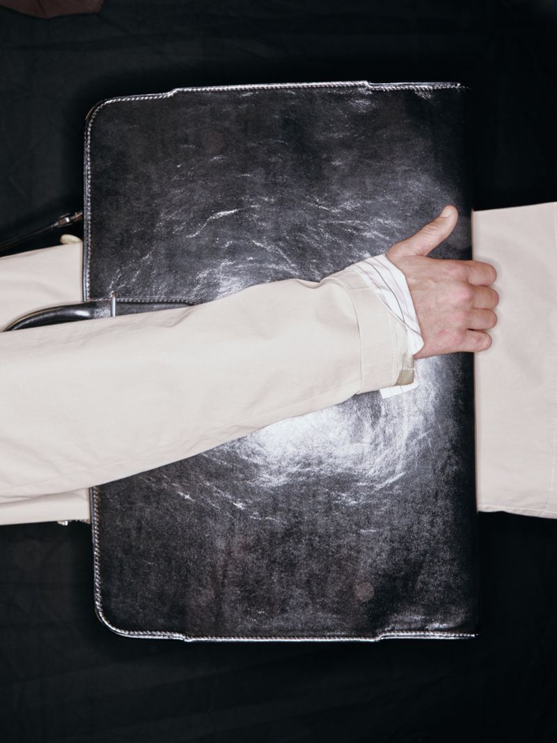 Overhead studio shot of a man's hand holding a closed, shiny black document briefcase against a dark backdrop.