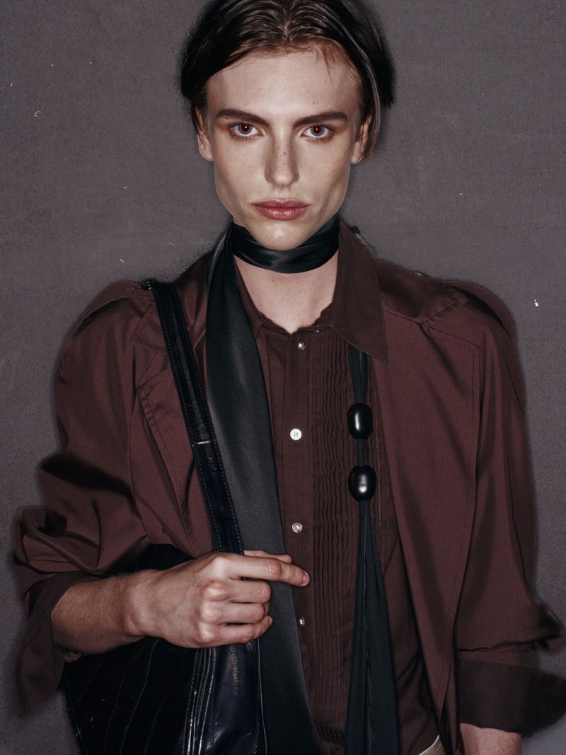 Studio portrait of a young adult male with fair skin, wearing a brown shirt, black scarf, and carrying a black bag over his shoulder.