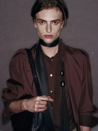 Studio portrait of a young adult male with fair skin, wearing a brown shirt, black scarf, and carrying a black bag over his shoulder.