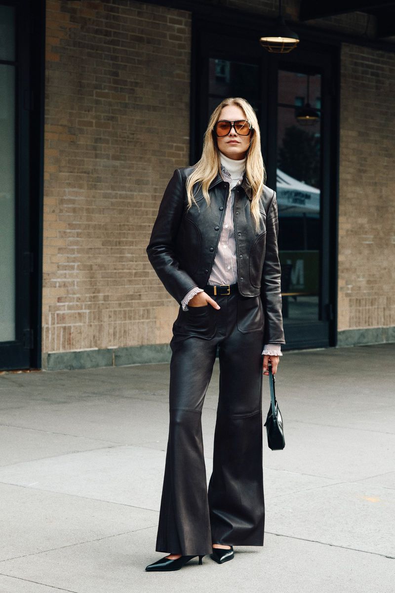 Fashion model in a dark leather jacket, wide-leg flared leather trousers, and oversized tinted sunglasses standing on a paved sidewalk.