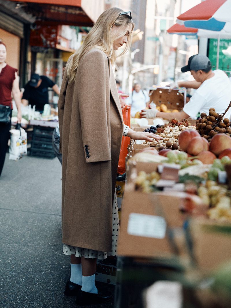 A woman in a tan coat browses fresh produce at an outdoor market on a sunny day. A vendor stands behind her, arranging the fruit.