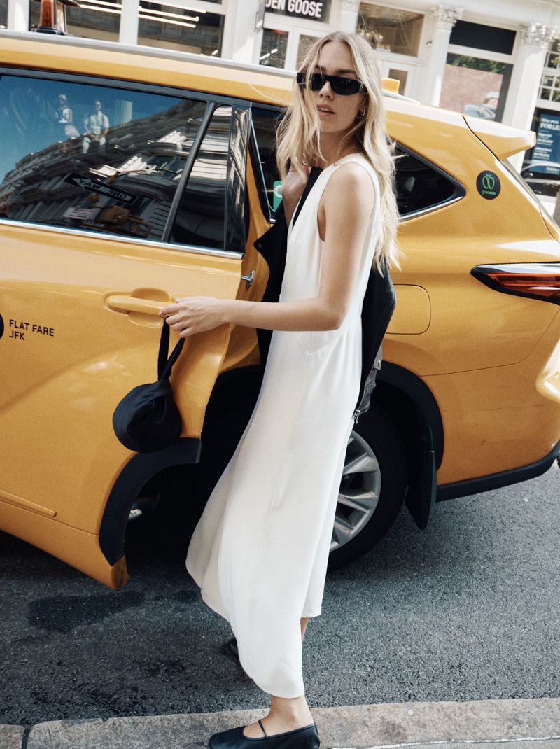 A blonde woman in a white dress exits a yellow taxi in an urban setting, wearing sunglasses and holding a black jacket.