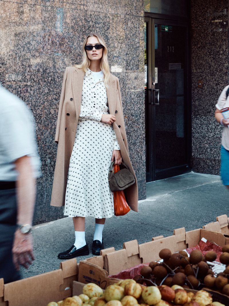 A woman in a polka dot dress and tan coat stands on a city street next to a fruit stand, with people passing by in the background.
