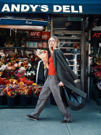 Woman in coat carrying flowers and purse walking past Andy's Deli storefront with vibrant floral displays on city sidewalk.