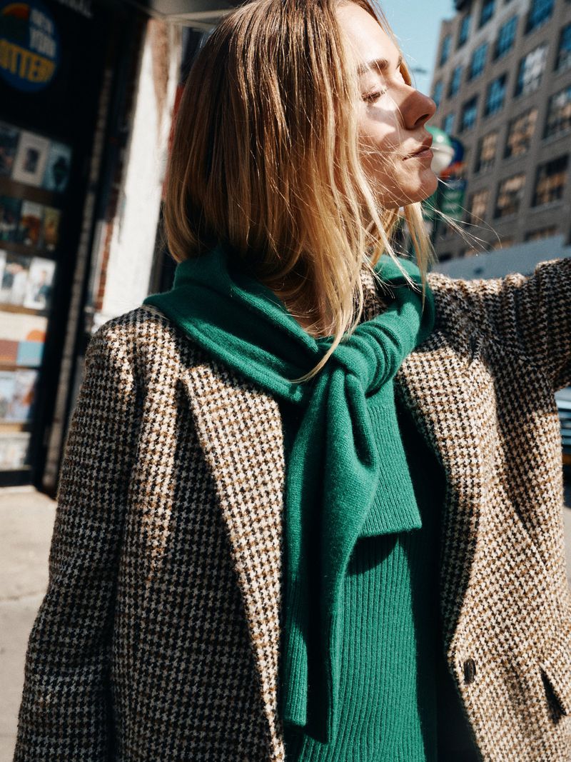 Woman in houndstooth blazer and green scarf stands in sunlight against urban backdrop, showcasing street style fashion outdoors.