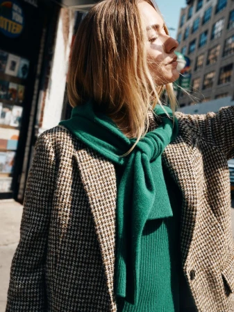 Woman in houndstooth blazer and green scarf stands in sunlight against urban backdrop, showcasing street style fashion outdoors.