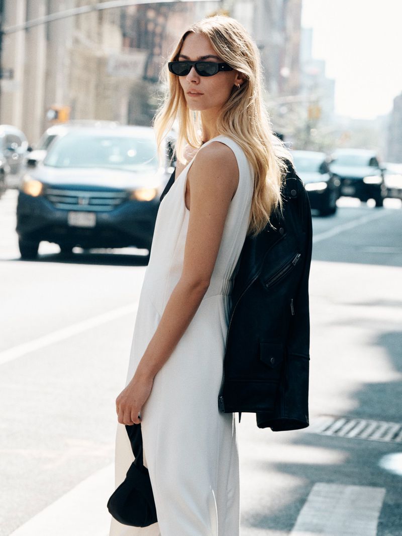 Blonde woman wearing a sleeveless white dress and black sunglasses with a black leather jacket draped over her shoulder on a city street.