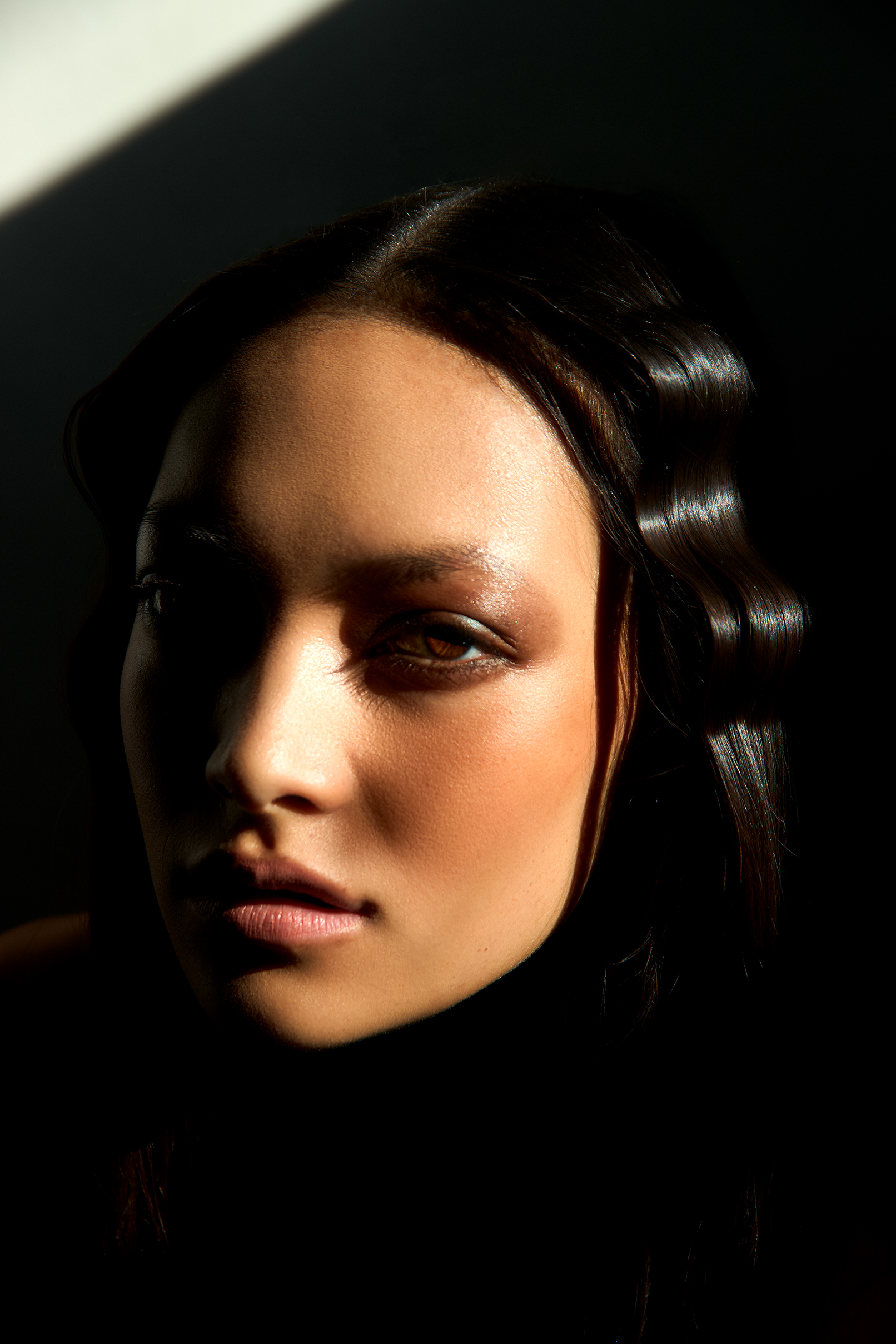 Close-up portrait of a woman with dark, wavy hair in dramatic lighting, showcasing her face and skin texture.