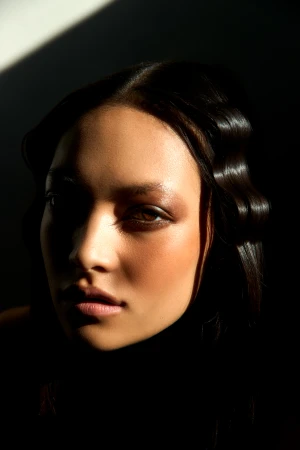 Close-up portrait of a woman with dark, wavy hair in dramatic lighting, showcasing her face and skin texture.