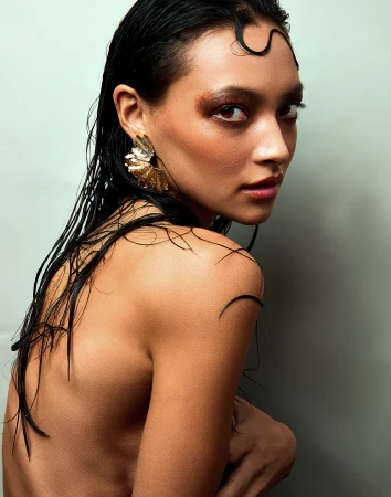 Close-up beauty portrait of a woman with wet hair across her face, iridescent eye makeup, and a textured gold earring.