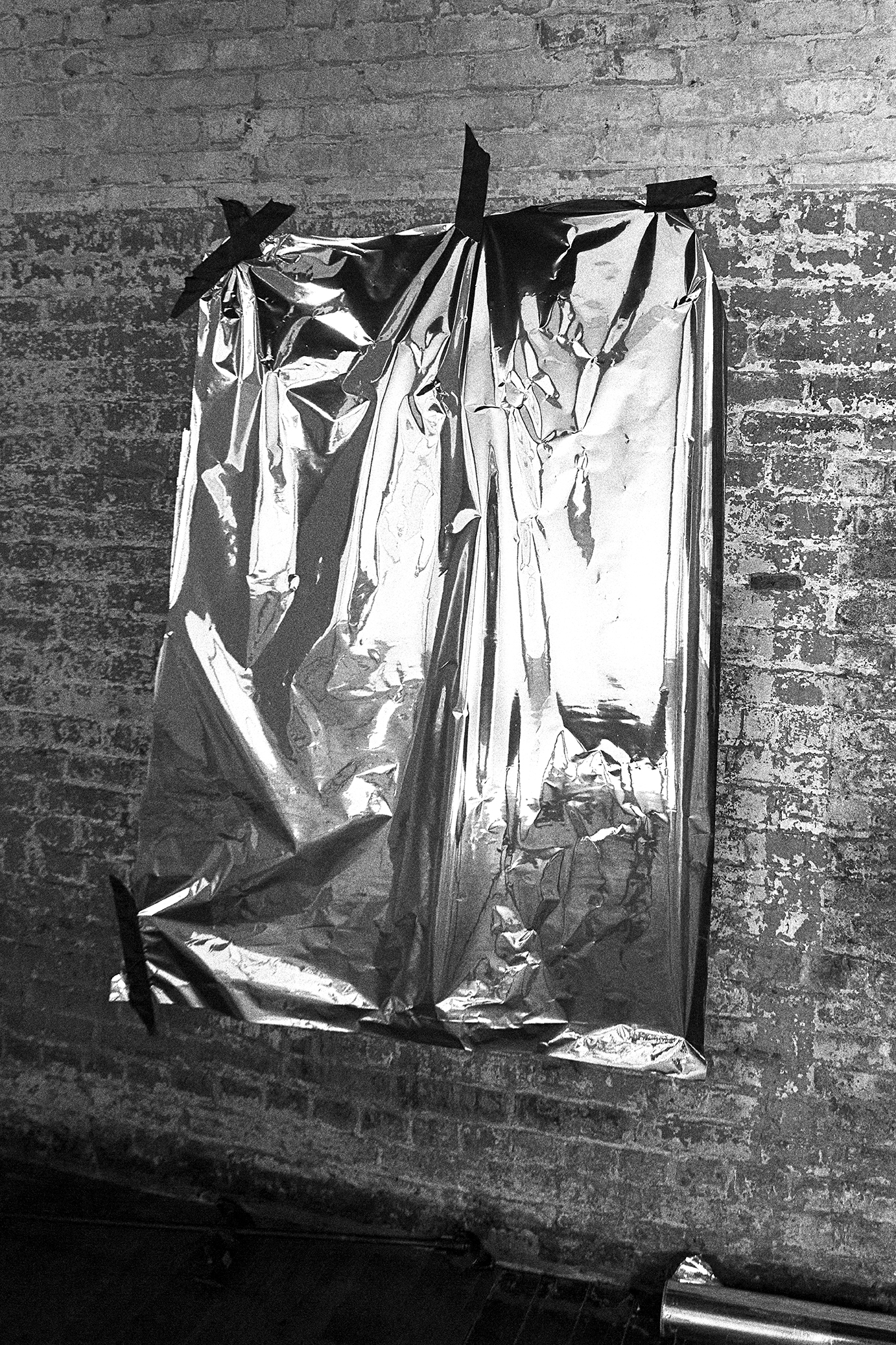 Monochrome image of a rectangular metallic sheet, secured with tape to a weathered brick wall indoors, creating a stark contrast.
