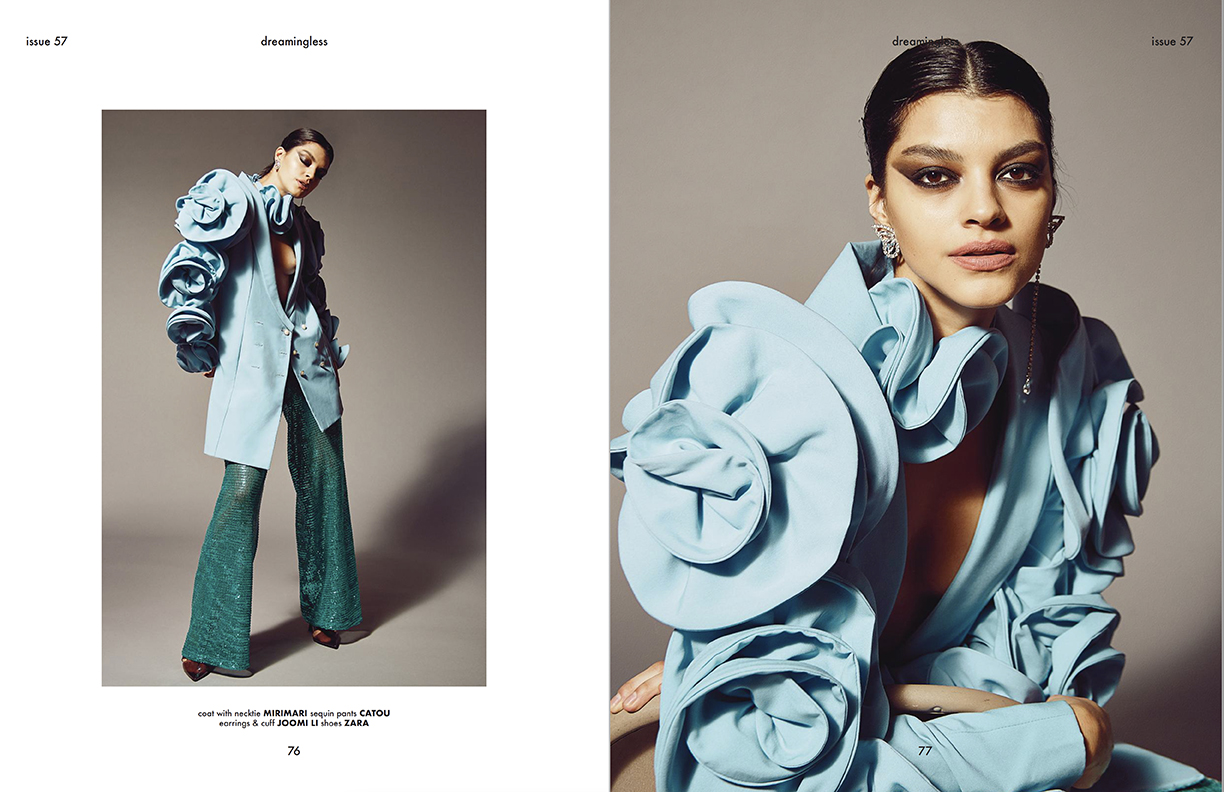Fashion editorial portrait of a woman in a statement blue coat with floral ruffles and green sequin pants against a gray background.