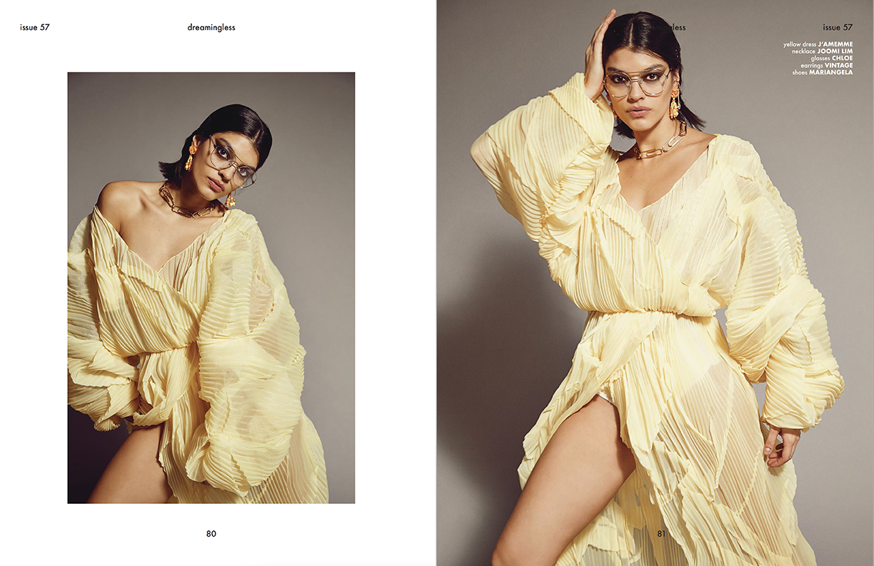 Brunette model in flowing yellow dress and retro glasses poses against a grey backdrop in a fashion editorial shot indoors.