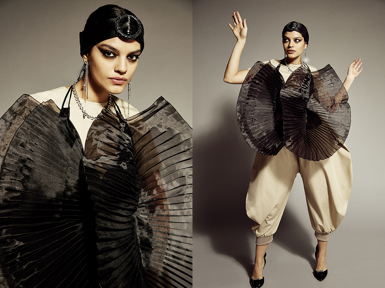 Fashionable adult female poses in a black pleated top, beige pants, black headpiece, and pumps against a neutral studio backdrop.