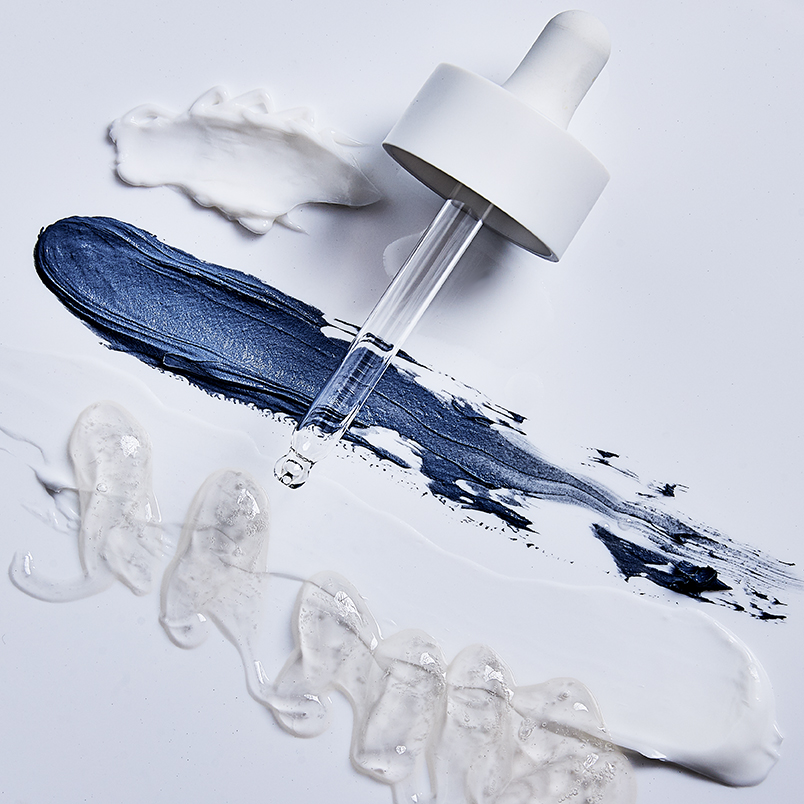Overhead shot of clear liquid serum in a dropper with smears of blue, white and clear cream on a white surface in studio lighting.