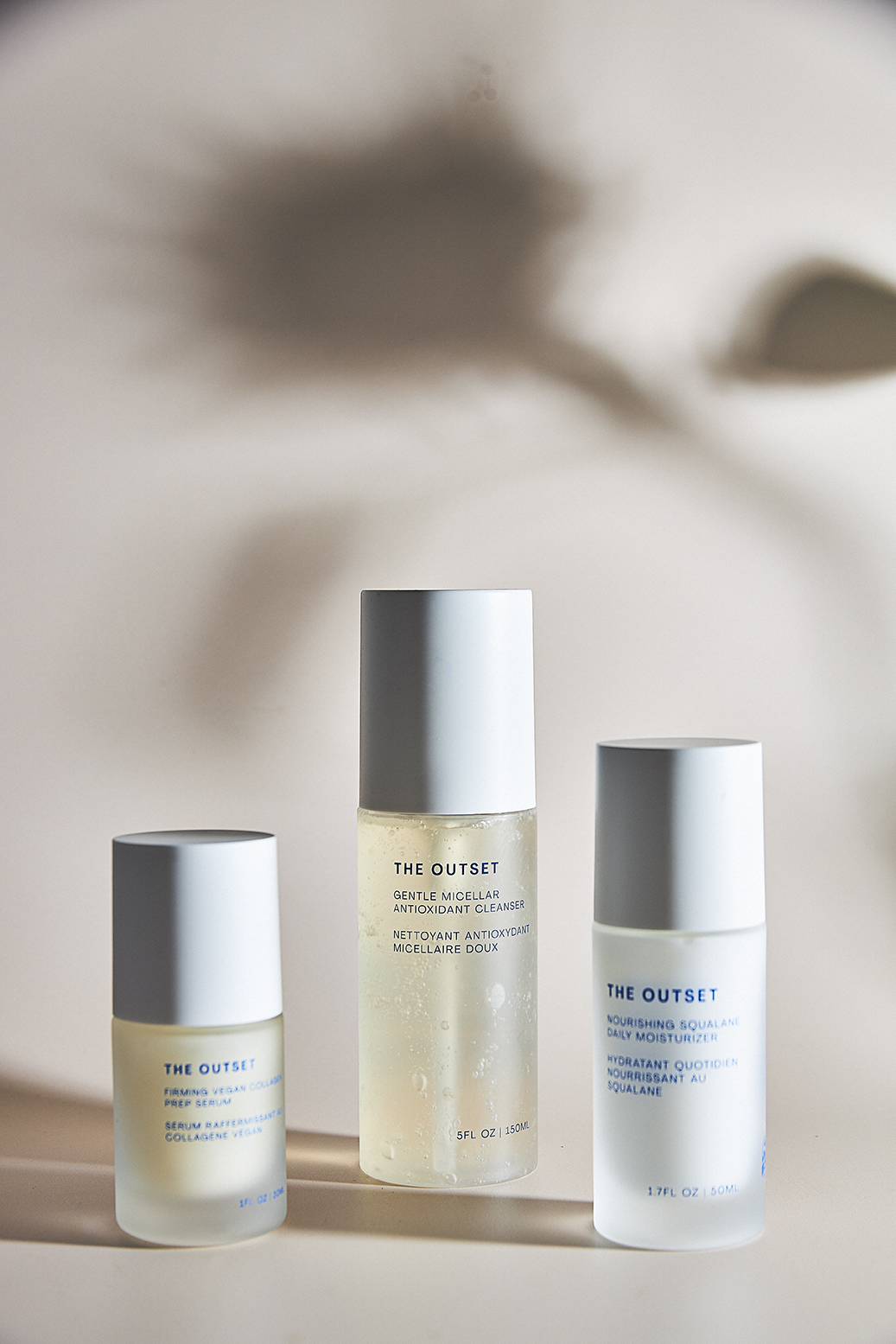 Three 'The Outset' branded skincare product bottles on a plain surface with plant shadow behind them. Beauty and skincare concept.