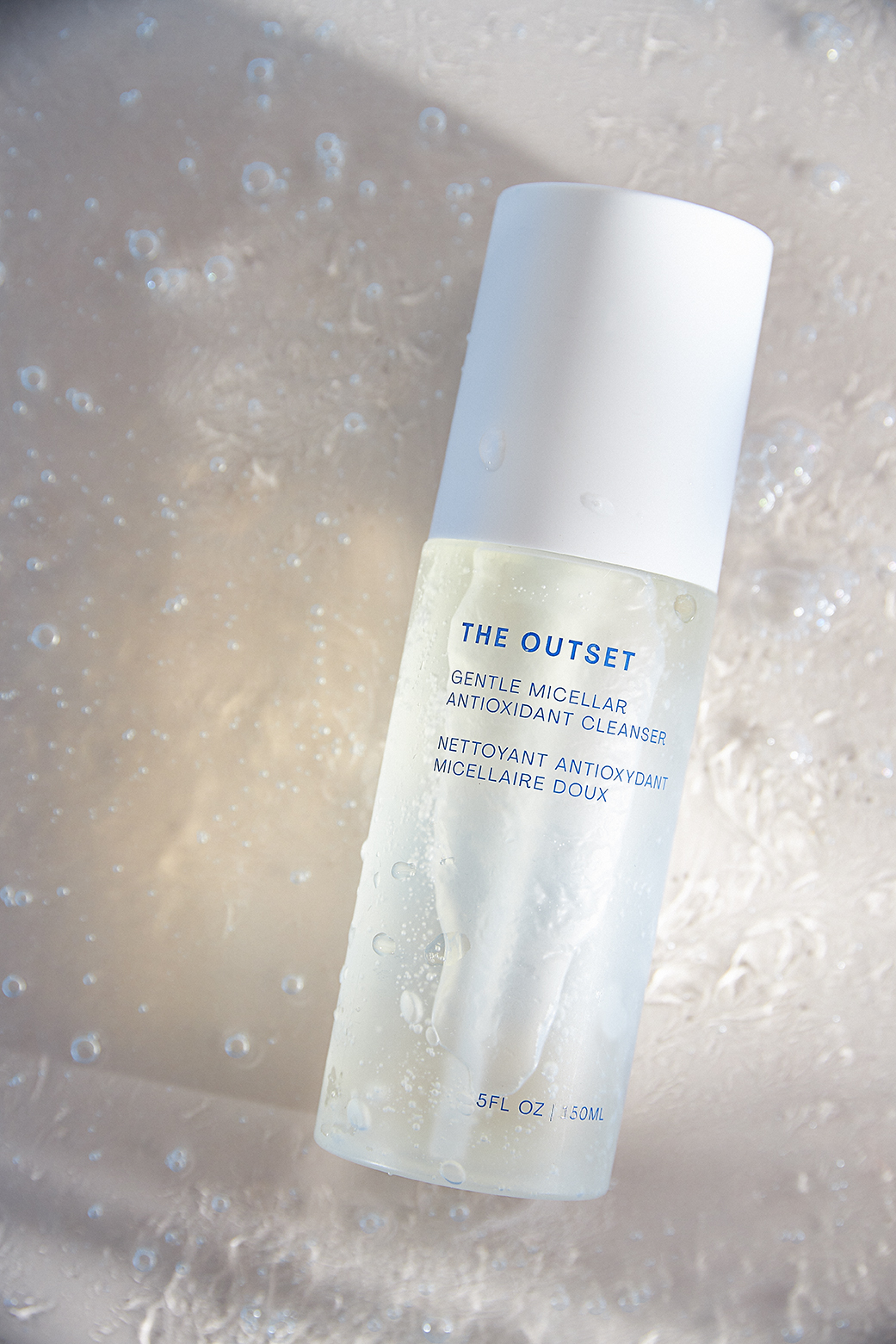 A white bottle of The Outset micellar antioxidant cleanser on a textured light background with water droplets on the bottle.