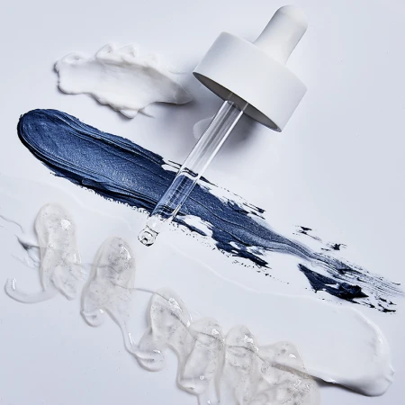 Glass cosmetic dropper placed over dark blue cream, white lotion, and clear gel swatches on a white background.