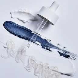 Glass cosmetic dropper placed over dark blue cream, white lotion, and clear gel swatches on a white background.