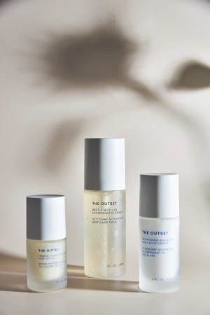 Three frosted glass skincare bottles with white caps from The Outset arranged against a warm neutral backdrop with floral shadows.