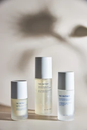 Three frosted glass skincare bottles with white caps from The Outset arranged against a warm neutral backdrop with floral shadows.