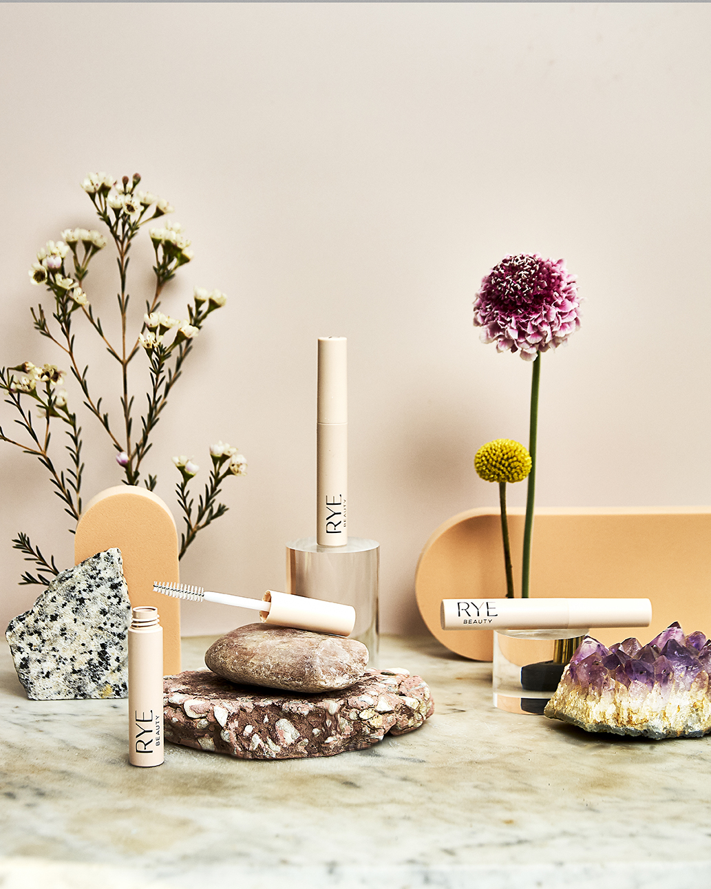 Still life of Rye Beauty mascara tubes arranged with flowers and stones in a minimalist studio setting, promoting cosmetics.