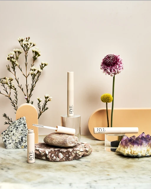 Still life of Rye Beauty mascara tubes arranged with flowers and stones in a minimalist studio setting, promoting cosmetics.