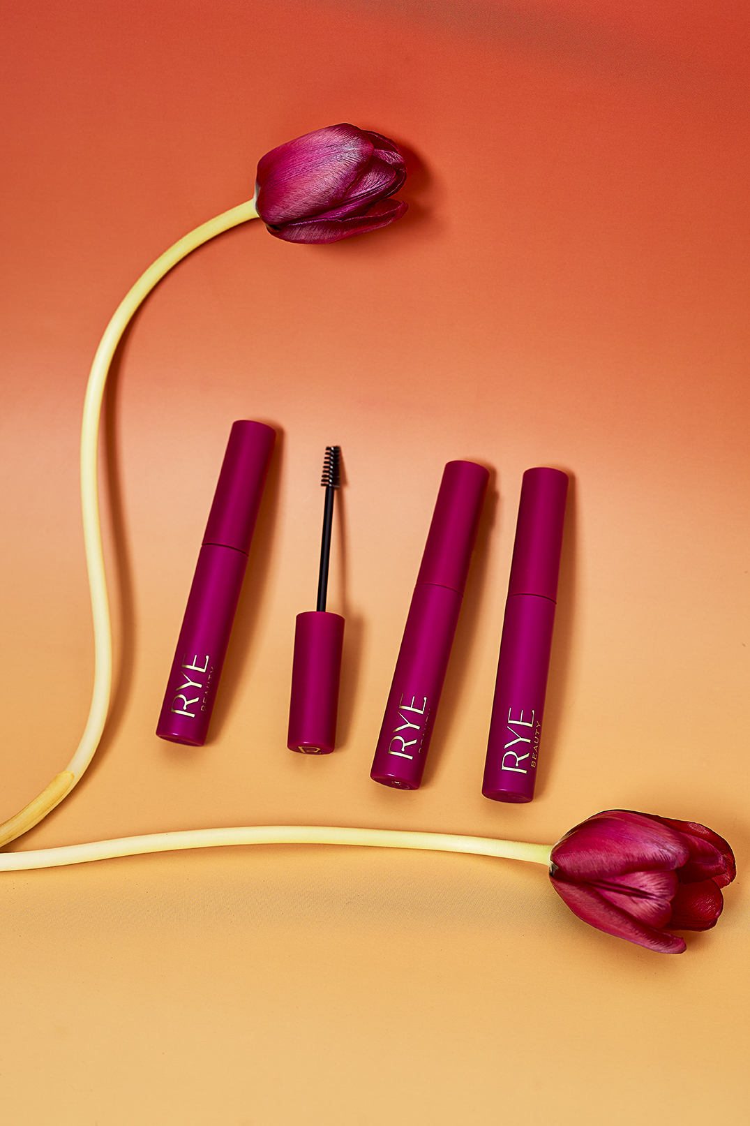 Product shot of three magenta mascara tubes from RYE with one applicator placed between them, flanked by two magenta tulips, on a peach background.
