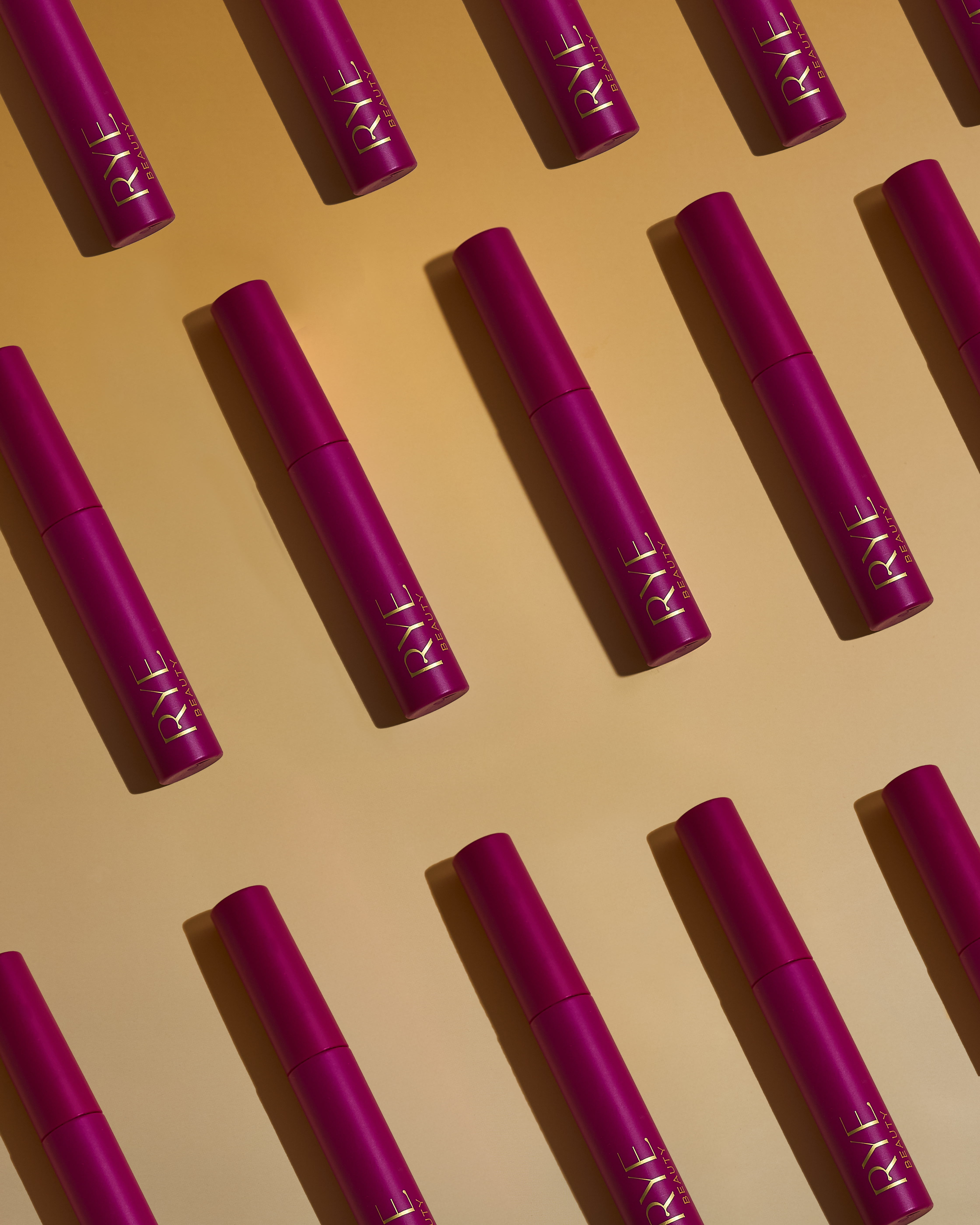 Overhead shot of multiple pink beauty product tubes with gold lettering, arranged in a diagonal pattern on a light orange background.