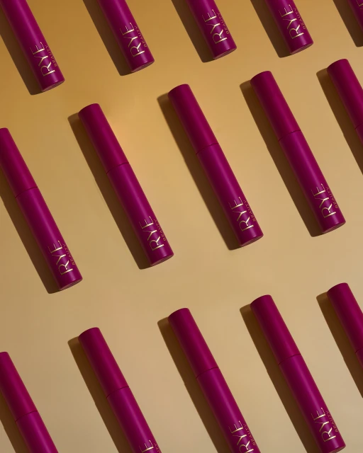 Overhead shot of multiple pink beauty product tubes with gold lettering, arranged in a diagonal pattern on a light orange background.