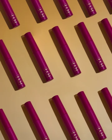 Overhead shot of multiple pink beauty product tubes with gold lettering, arranged in a diagonal pattern on a light orange background.